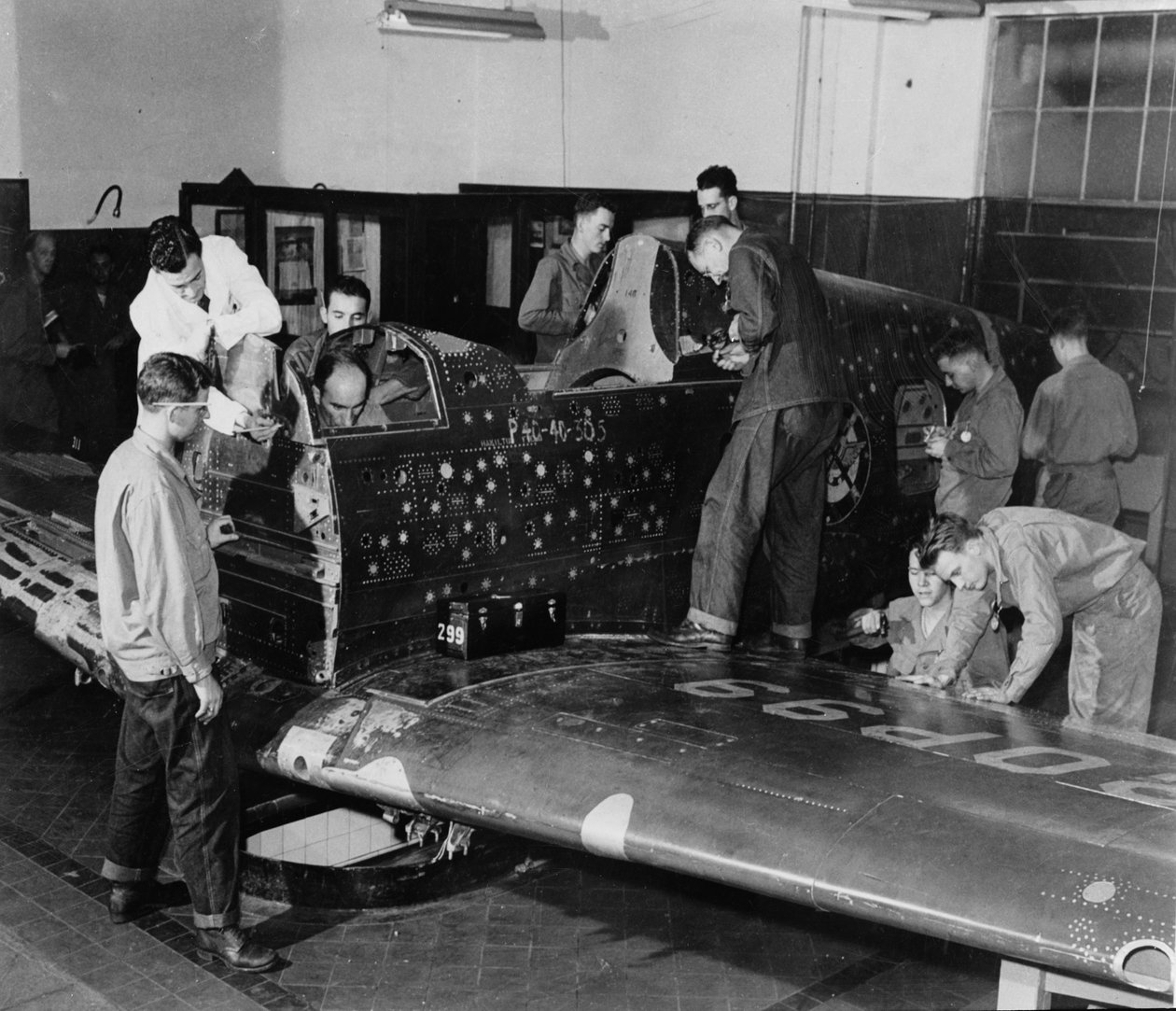 Asker stajyerleri, simüle edilmiş kurşun delikleri ve yamaları olan, motor veya kuyruk tertibatı olmayan bir Curtiss "Warhawk" savaş uçağı olan "Uçan Yama" üzerinde çalışıyor, c.1942 by American Photographer