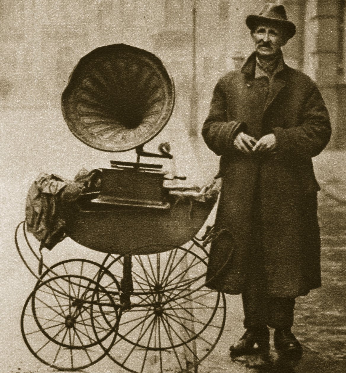 Bir sokak satıcısı, bir çocuk arabası içinde taşınan bir gramofon ile eğlendirir (sepya fotoğrafı) by English Photographer