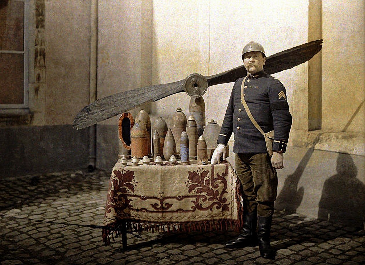 Alman el bombaları ve uçak pervanesi olan bir masanın yanında bir Fransız askeri, Reims, Marne, 1917 (otokrom) by Fernand Cuville
