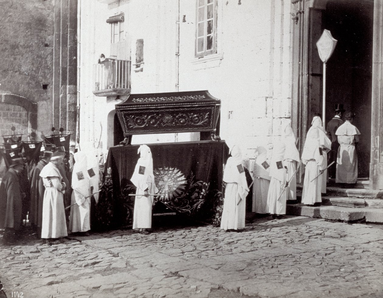 Napoli'deki San Francesco Cemaati'nin cenaze töreni. Ön planda, bir kilisenin önünde, beyaz tunikli ve kapüşonlu çok sayıda 'kardeş kardeş', üzerine tabutun yerleştirildiği işlemeli bir bezle süslenmiş bir katafalka eşlik ediyor. by Giorgio Sommer