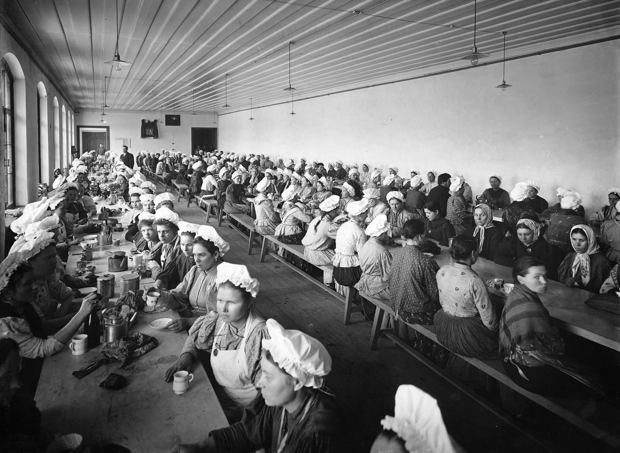 Bir fabrika deposundaki kadın işçiler, birkaç erkek gözetmenin gözetiminde yemek molası veriyor by Russian Photographer