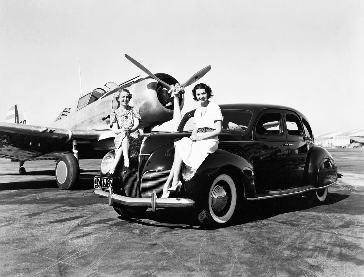 California: 1939 İki kadın, arkalarında tek motorlu bir uçak park etmiş bir havaalanında 1939 model Lincoln Zephyr sedanın çamurluklarında oturuyor (s/b fotoğraf) by Unbekannt
