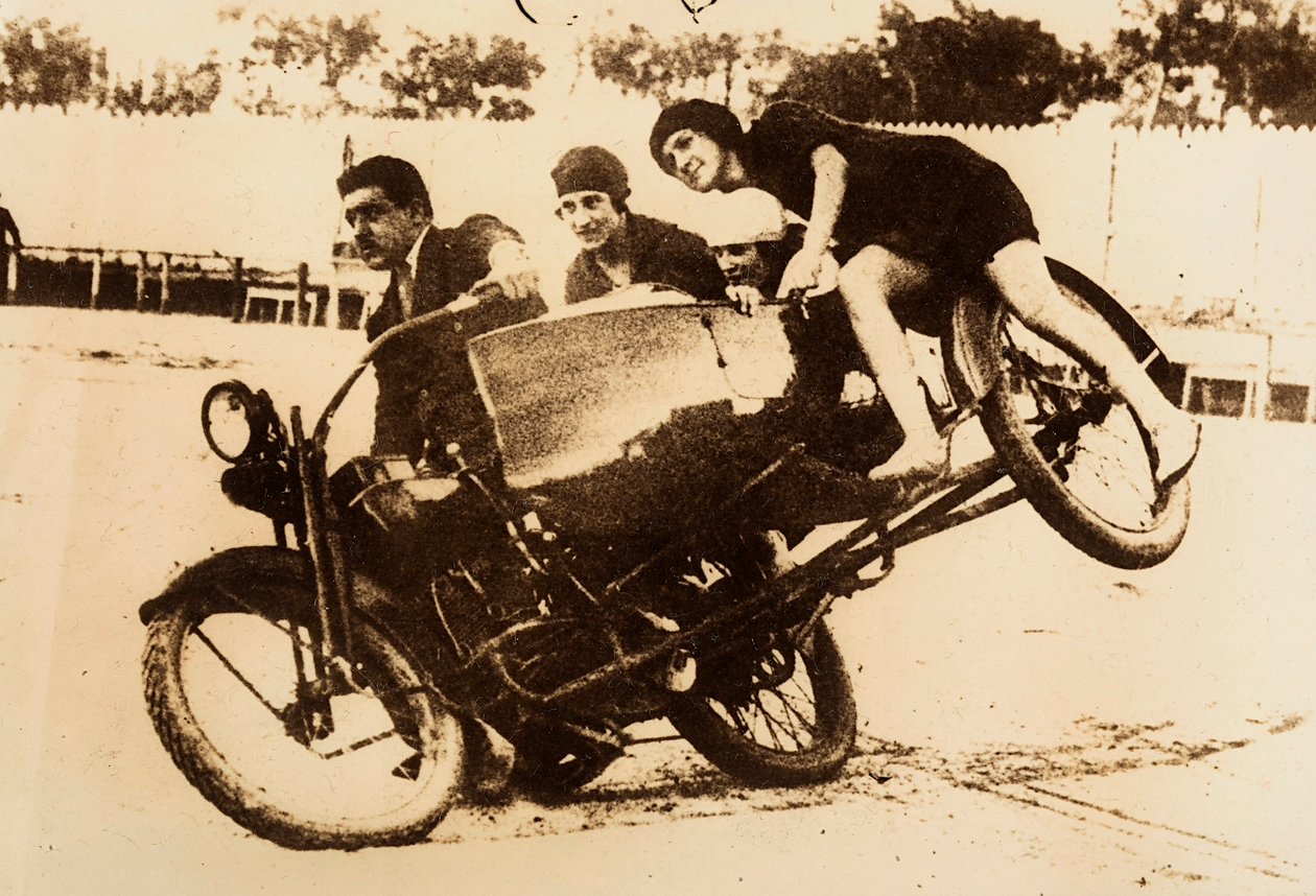 Bir adam üç genç kadını taşıyan sepetli bir motosikleti sürerek şov yapıyor. Dönüş yaptıklarında sepet yerden kalkıyor. by Unknown photographer