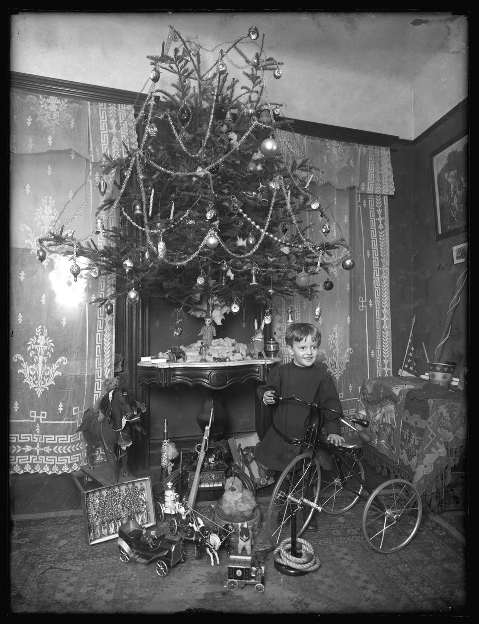 Seymour çocuğu salonda Noel ağacının yanında üç tekerlekli bisikletle poz verdi, Noel 1912 by William Davis Hassler