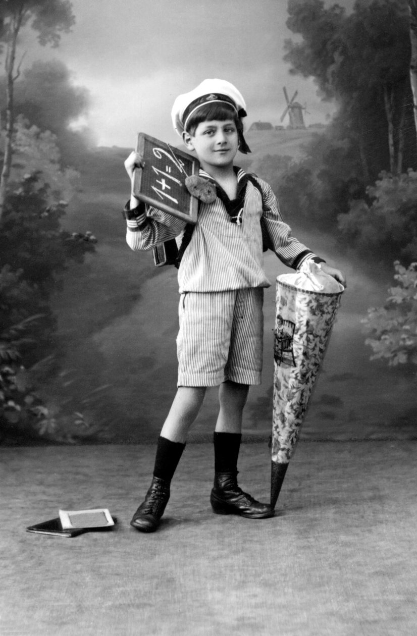 Okulun ilk günü. Üniformalı genç bir çocuk, 1+1 = 2 ekini gösteren bir levha ve sürpriz bir kese ile poz veriyor. 1910 civarında fotoğraf. by Ferdinand Georg Waldmuller