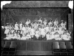 Küçük bir grup kız ellerinde oyuncak bebeklerle küçük bir sahnede poz verdi, muhtemelen Roma Katolik Yetim Sığınağı, Kingsbridge, Bronx, 1911 dolayları