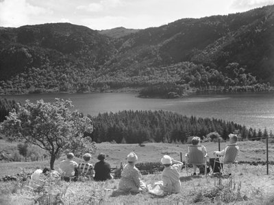 Çimlerde veya şezlonglarda oturmuş Thirlmere manzarasına bakan bir grup insan by Joseph Hardman